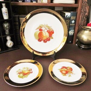 May include: Three white ceramic plates with gold trim, each featuring a different fruit design. The centre plate has a design of red apples, pears, and grapes. The two smaller plates have a design of red apples and grapes.
