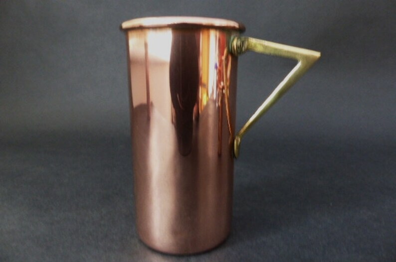 May include: A copper mug with a brass handle. The mug is cylindrical and has a smooth, polished surface.