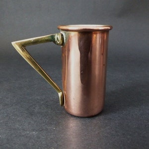 May include: A copper measuring cup with a brass handle. The cup is shiny and has a rounded bottom. The handle is attached to the side of the cup and has a triangular shape.