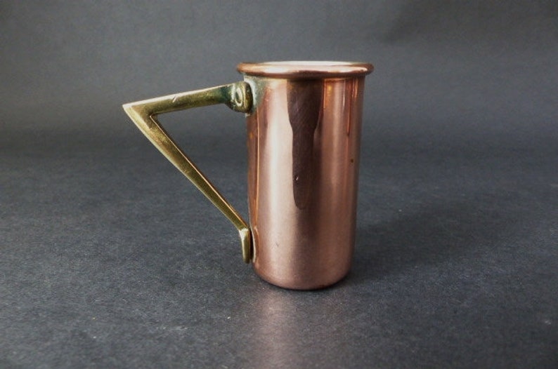 May include: A copper measuring cup with a brass handle. The cup is shiny and has a rounded bottom. The handle is attached to the side of the cup and has a triangular shape.