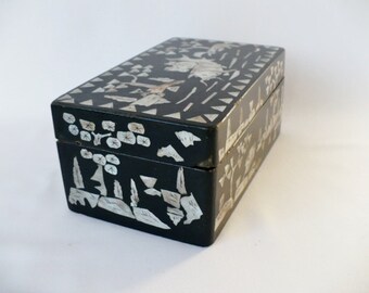Może przedstawiać: Czarne drewniane pudełko z ozdobnym wzorem z białej perłowej intarsji. Pudełko jest prostokątne i ma pokrywkę na zawiasach.