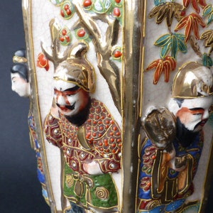 Może przedstawiać: Biały ceramiczny wazon z pozłacanym wykończeniem i misternymi ręcznie malowanymi detalami. Wazon przedstawia scenę z japońskimi postaciami w tradycyjnych strojach, z drzewem i kwiatami w tle. Postacie są namalowane w żywych kolorach, w tym czerwonym, niebieskim, zielonym i złotym.