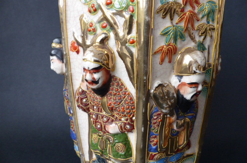 Może przedstawiać: Biały ceramiczny wazon z pozłacanym wykończeniem i misternymi ręcznie malowanymi detalami. Wazon przedstawia scenę z japońskimi postaciami w tradycyjnych strojach, z drzewem i kwiatami w tle. Postacie są namalowane w żywych kolorach, w tym czerwonym, niebieskim, zielonym i złotym.