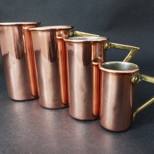 May include: Set of five copper measuring cups with brass handles. The cups are graduated in size, with the largest cup being the tallest. The cups are all polished to a shine.