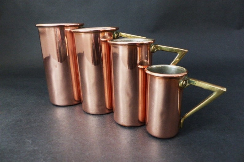 May include: Set of five copper measuring cups with brass handles. The cups are graduated in size, with the largest cup being the tallest. The cups are all polished to a shine.