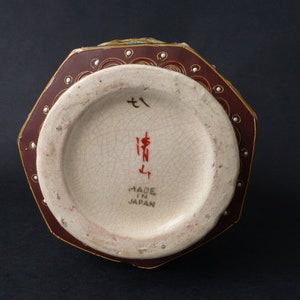 Może przedstawiać: Ośmiokątny wazon ceramiczny z czerwono-złotym wzorem. Podstawa wazonu ma białą glazurę z japońskimi znakami i napisem "Made in Japan".