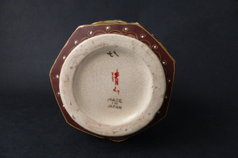 Może przedstawiać: Ośmiokątny wazon ceramiczny z czerwono-złotym wzorem. Podstawa wazonu ma białą glazurę z japońskimi znakami i napisem "Made in Japan".