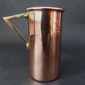 May include: A copper jug with a brass handle. The jug is cylindrical in shape and has a smooth, polished surface.