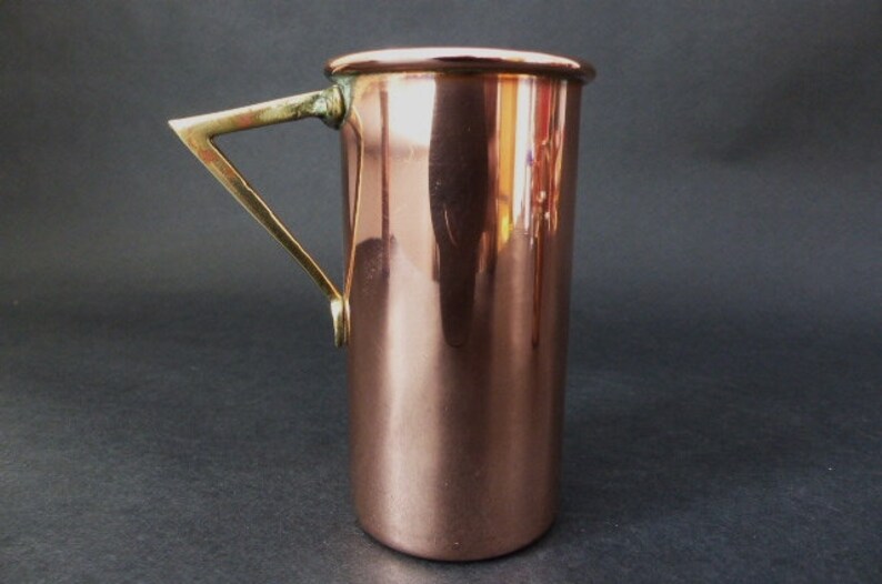 May include: A copper jug with a brass handle. The jug is cylindrical in shape and has a smooth, polished surface.