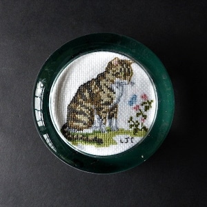 May include: A green glass paperweight featuring a white embroidered image of a tabby cat sitting with a butterfly and flowers. The embroidery is signed "LJ."
