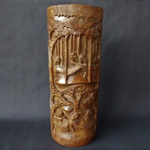 May include: A tall, cylindrical wooden vase with intricate carvings depicting a scene of people and trees. The vase is made of dark brown wood and has a smooth, polished finish.