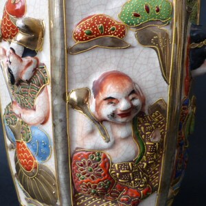 Może przedstawiać: Biały ceramiczny wazon o sześciokątnym kształcie, ozdobiony kolorowymi postaciami malowanymi i złotymi akcentami. Wazon przedstawia scenę z wieloma postaciami, w tym mężczyzną w czerwonym kapeluszu, kobietą o czarnych włosach i mężczyzną z czerwoną brodą. Wazon jest w tradycyjnym japońskim stylu.