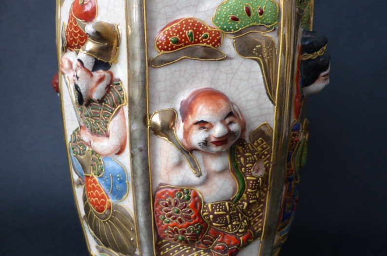 Może przedstawiać: Biały ceramiczny wazon o sześciokątnym kształcie, ozdobiony kolorowymi postaciami malowanymi i złotymi akcentami. Wazon przedstawia scenę z wieloma postaciami, w tym mężczyzną w czerwonym kapeluszu, kobietą o czarnych włosach i mężczyzną z czerwoną brodą. Wazon jest w tradycyjnym japońskim stylu.