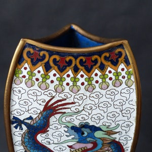 K&ouml;nnte beinhalten: Eine wei&szlig;e Keramikvase mit einem blauen und roten Drachenmuster. Die Vase hat einen goldenen Rand und ein dekoratives Muster um die Oberseite.
