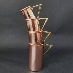 May include: Set of four copper mugs with brass handles. The mugs are stacked on top of each other, with the handles forming a triangular shape.