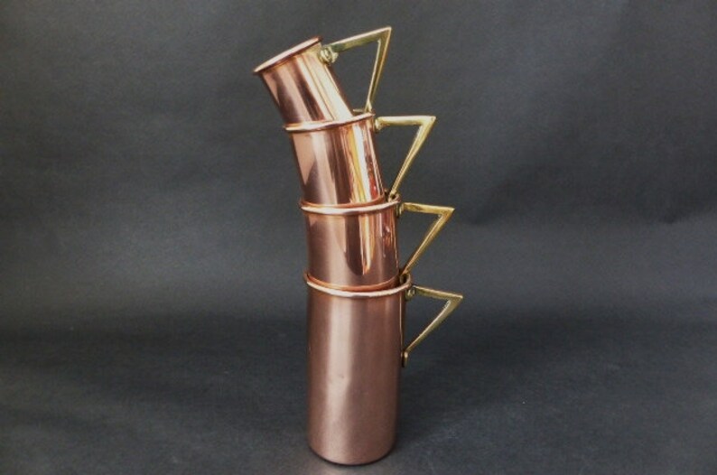 May include: Set of four copper mugs with brass handles. The mugs are stacked on top of each other, with the handles forming a triangular shape.