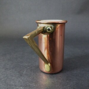 May include: A small copper measuring cup with a brass handle. The cup has a white enamel interior.