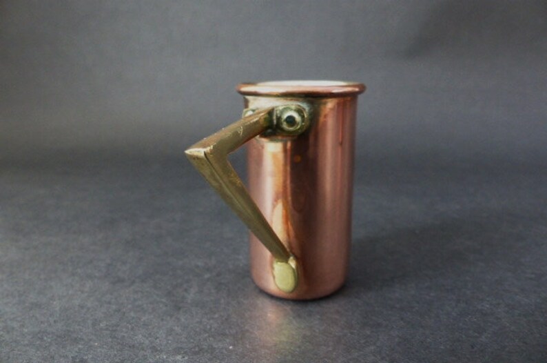 May include: A small copper measuring cup with a brass handle. The cup has a white enamel interior.