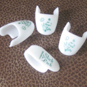 May include: Four white finger picks with green graphics and text. One pick has the text "Slick Pick". The other picks have musical notes and the letters "D" and "1". The picks are on a brown textured surface.