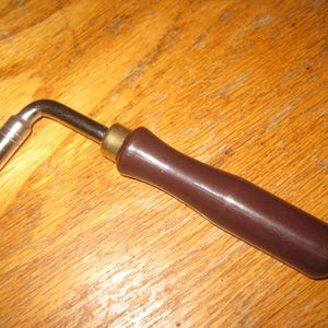 May include: A brown handled tuning key for a stringed instrument. The key is made of metal and has a L-shaped handle.