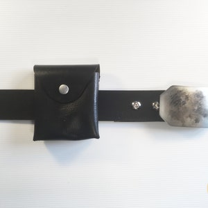 May include: A black leather belt with a small black leather pouch and a silver metal buckle. The pouch has a flap with a silver button.