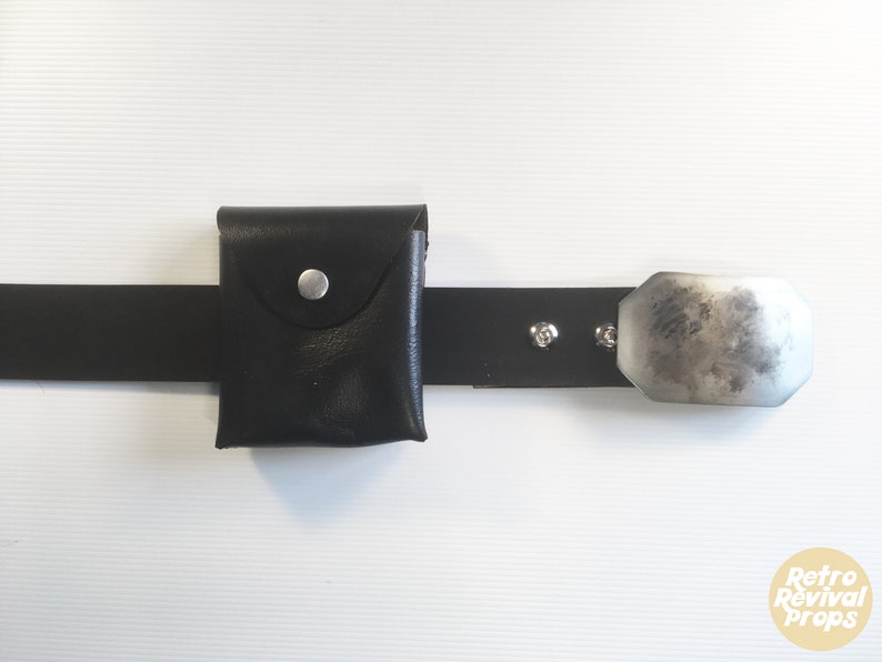 May include: A black leather belt with a small black leather pouch and a silver metal buckle. The pouch has a flap with a silver button.