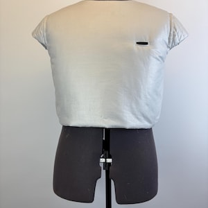May include: A light silver-colored, short-sleeved garment with a small black rectangular detail, displayed on a black dress form. The garment has a quilted texture on the sleeves.