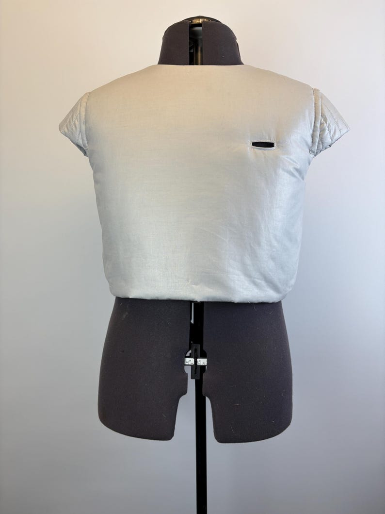 May include: A light silver-colored, short-sleeved garment with a small black rectangular detail, displayed on a black dress form. The garment has a quilted texture on the sleeves.