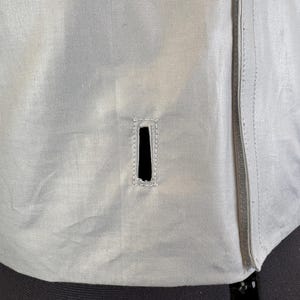 May include: Close-up of a light gray jacket with a vertical zipper on the right side. The jacket has a rectangular vent with reinforced stitching. The fabric appears to be a lightweight material.
