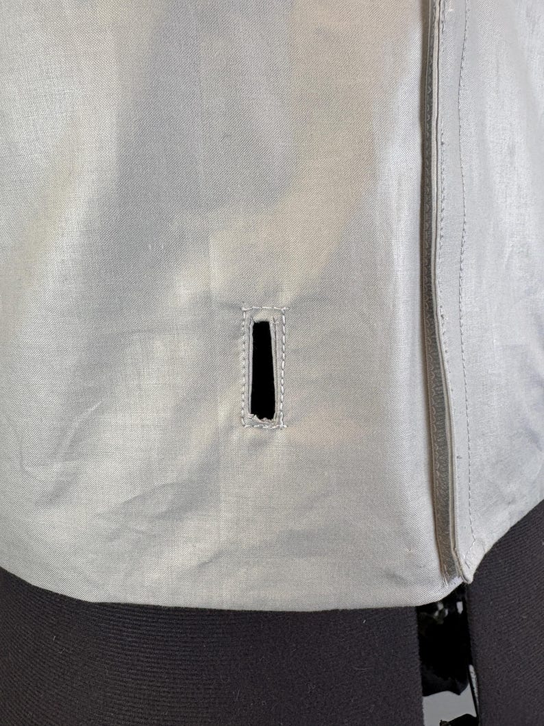 May include: Close-up of a light gray jacket with a vertical zipper on the right side. The jacket has a rectangular vent with reinforced stitching. The fabric appears to be a lightweight material.