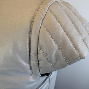 May include: Close-up of a light beige garment's shoulder and sleeve. The sleeve features a quilted design with parallel stitching. The fabric appears smooth and the stitching is in a lighter shade, creating a subtle contrast.