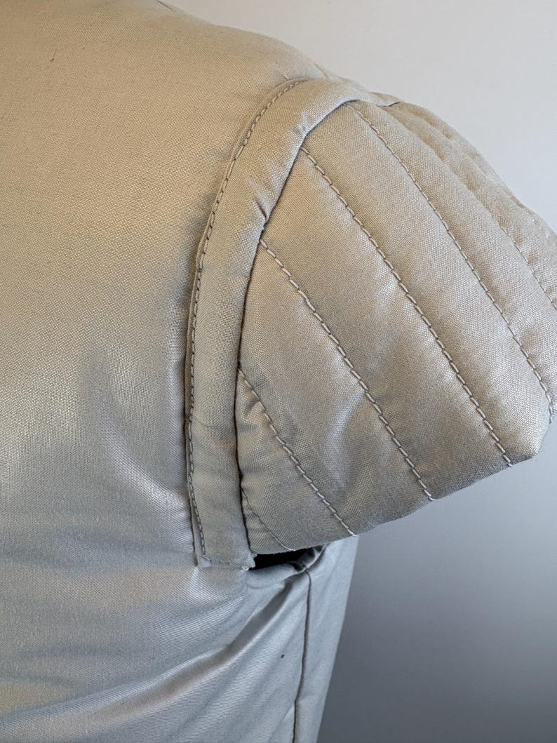 May include: Close-up of a light beige garment's shoulder and sleeve. The sleeve features a quilted design with parallel stitching. The fabric appears smooth and the stitching is in a lighter shade, creating a subtle contrast.
