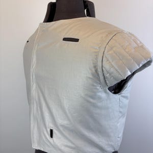 May include: A light gray, short-sleeved garment with a quilted shoulder detail and a small rectangular opening. The garment is displayed on a black dress form. The fabric has a slight sheen.