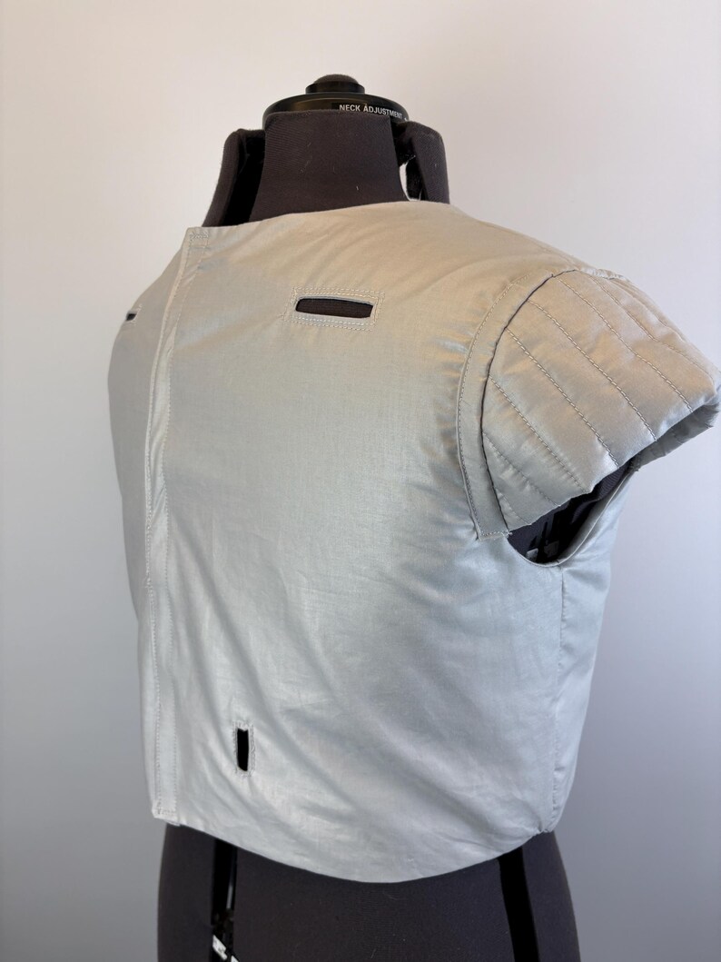 May include: A light gray, short-sleeved garment with a quilted shoulder detail and a small rectangular opening. The garment is displayed on a black dress form. The fabric has a slight sheen.