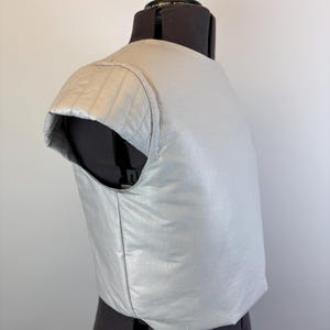 May include: A light gray, padded garment, possibly a vest or part of a costume, is displayed on a black dress form. The garment has a quilted shoulder detail and a smooth, shiny texture. The design suggests a focus on protection or padding.