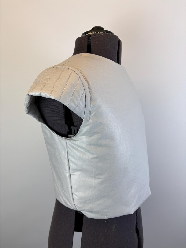 May include: A light gray, padded garment, possibly a vest or part of a costume, is displayed on a black dress form. The garment has a quilted shoulder detail and a smooth, shiny texture. The design suggests a focus on protection or padding.
