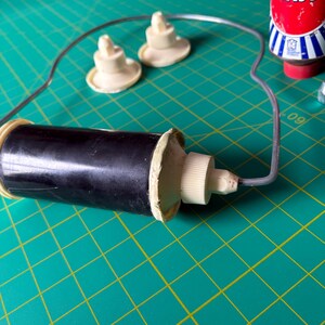 May include: A black cylindrical tool with a white threaded cap and a thin grey wire frame attached to the end. Two small, round, off-white plastic components with central pegs are positioned nearby, alongside a red bottle labeled BRASSO.