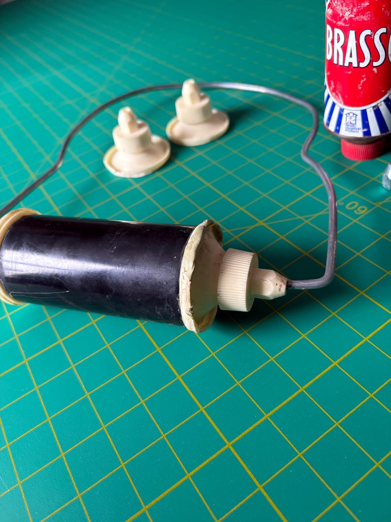 May include: A black cylindrical tool with a white threaded cap and a thin grey wire frame attached to the end. Two small, round, off-white plastic components with central pegs are positioned nearby, alongside a red bottle labeled BRASSO.