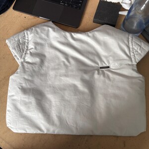 May include: A light gray vest-like garment with short sleeves, possibly a prototype or sample. The vest has a quilted shoulder detail and a small rectangular pocket. It is lying on a light brown surface, with a laptop and other items in the background.