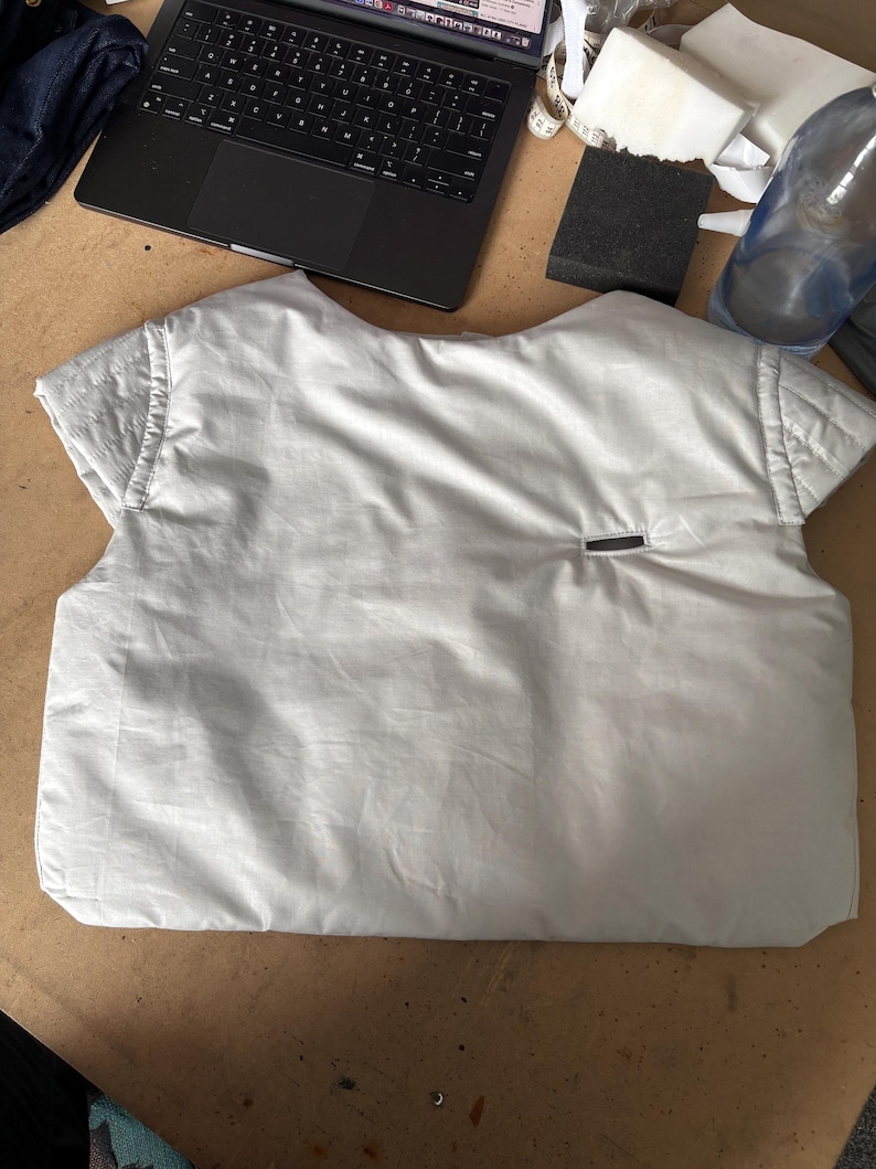 May include: A light gray vest-like garment with short sleeves, possibly a prototype or sample. The vest has a quilted shoulder detail and a small rectangular pocket. It is lying on a light brown surface, with a laptop and other items in the background.