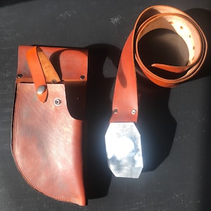May include: A brown leather belt with a silver buckle and a matching brown leather holster. The holster has a flap with a snap closure.