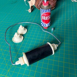 May include: A black cylindrical component with cream-colored end caps connected by a thin metal wire loop, two additional cream-colored circular parts, a small metal nut, and a metal can of Brasso polish with a red cap and blue and white stripes.