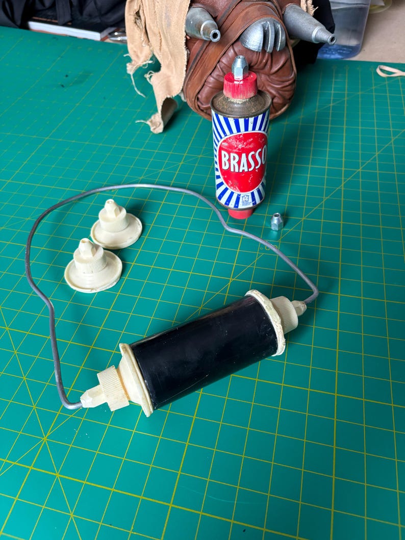 May include: A black cylindrical component with cream-colored end caps connected by a thin metal wire loop, two additional cream-colored circular parts, a small metal nut, and a metal can of Brasso polish with a red cap and blue and white stripes.