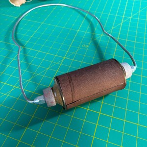 May include: A cylindrical brown leather-wrapped tool featuring a silver-toned metal wire handle attached to both ends. The central component has textured silver metal caps on each side, connected by a thin, curved silver wire frame.