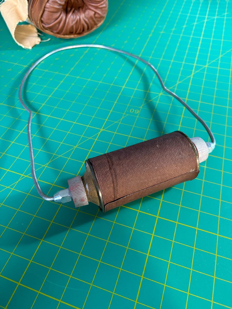 May include: A cylindrical brown leather-wrapped tool featuring a silver-toned metal wire handle attached to both ends. The central component has textured silver metal caps on each side, connected by a thin, curved silver wire frame.
