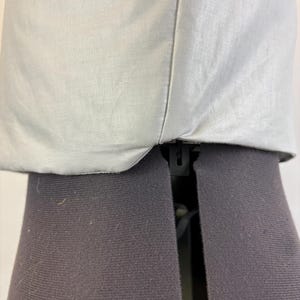 May include: Close-up of a light gray fabric garment. The fabric appears to be a lightweight material, possibly linen or cotton, with a subtle texture. The garment is sewn with a visible seam. The garment is placed on a black dress form.