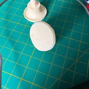 May include: Two off-white plastic components are visible. One is a circular base with a central nozzle tip, and the other is a smooth, oval-shaped piece. Both items are positioned on a green surface with a yellow grid pattern.