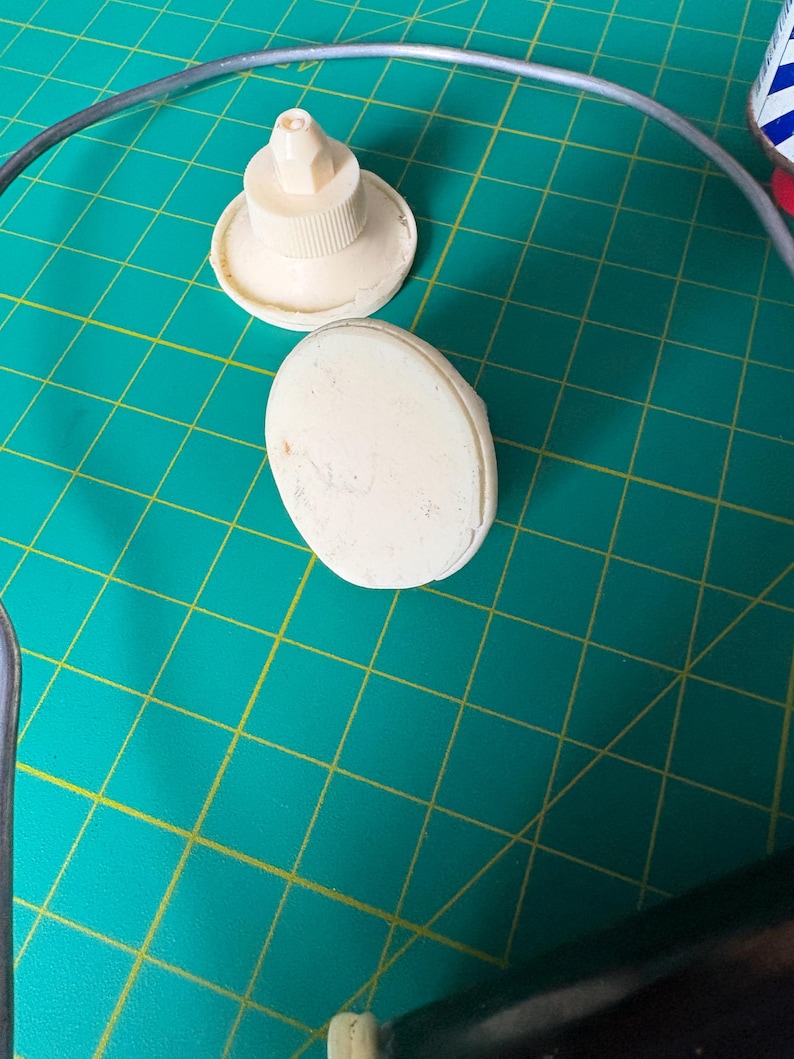 May include: Two off-white plastic components are visible. One is a circular base with a central nozzle tip, and the other is a smooth, oval-shaped piece. Both items are positioned on a green surface with a yellow grid pattern.