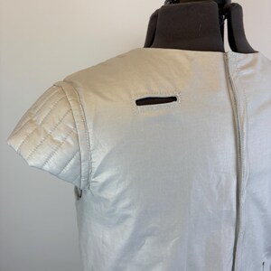 May include: A light grey, sleeveless jacket with a quilted shoulder detail and a zipper closure. The jacket has a small rectangular opening near the shoulder and a similar opening near the bottom. The garment is displayed on a mannequin.