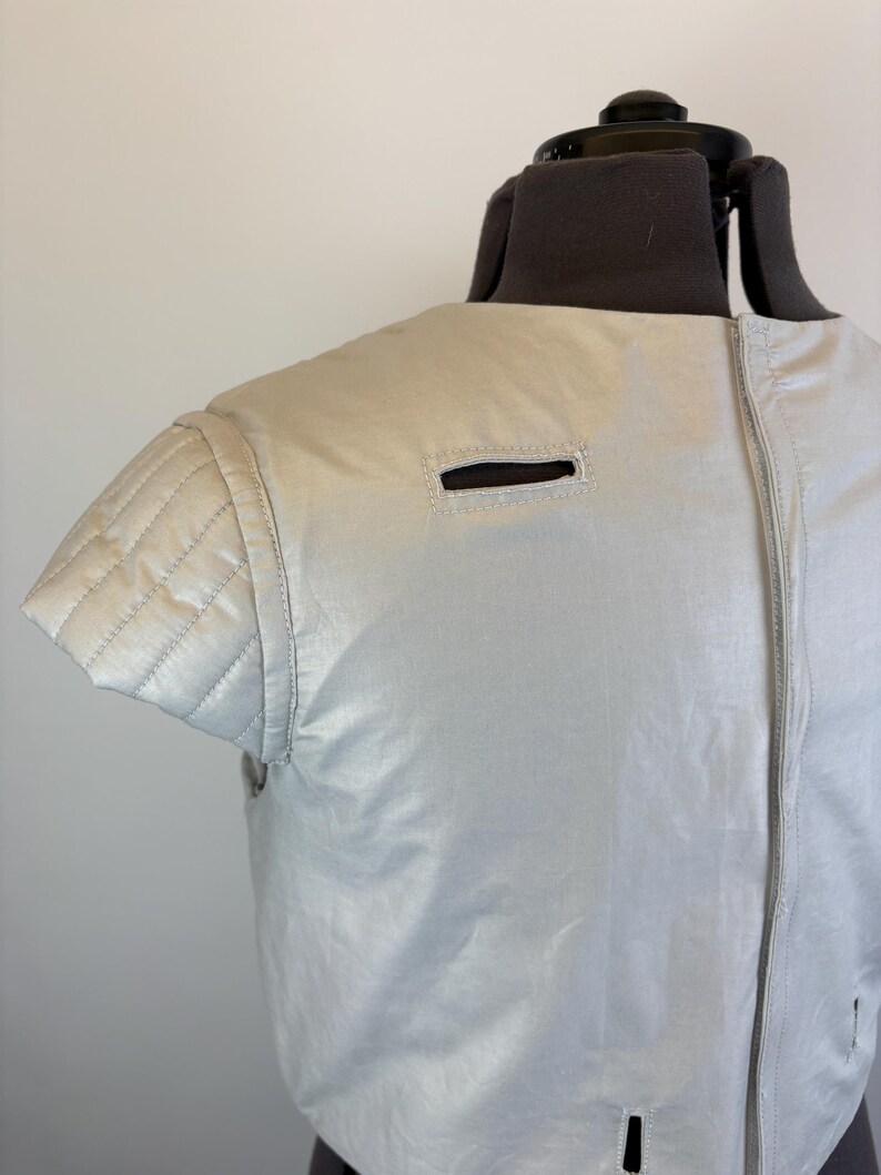 May include: A light grey, sleeveless jacket with a quilted shoulder detail and a zipper closure. The jacket has a small rectangular opening near the shoulder and a similar opening near the bottom. The garment is displayed on a mannequin.